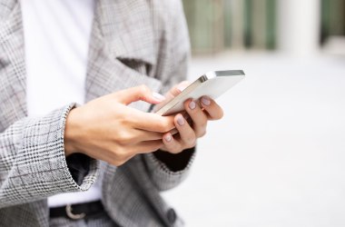 Online Services, © istockphoto/Prostock-Studio Hände einer Person mit hell lackierten Fingernägeln, die ein weißes Smartphone halten, bekleidet mit grauem Karoblazer und weißem Shirt.