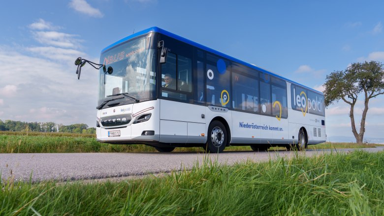 LEOpoldi Regionalbus in Niederösterreich, © NÖVOG/Bollwein Blauer und weißer Linienbus mit Aufschrift 'leo' und 'leo mobil' fährt auf Landstraße, Wiese und Baum im Hintergrund.
