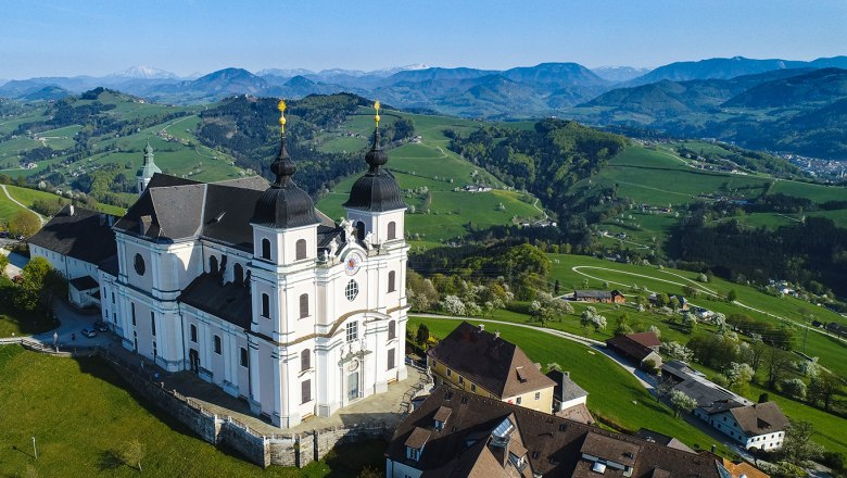 Blick ins Mostviertel, © Mostviertel Tourismus/Cleanhill Studios, Gerald Prüller Barocke Kirche mit zwei Türmen auf einem Hügel, umgeben von Häusern und grünen Wiesen, im Hintergrund Berge unter blauem Himmel.