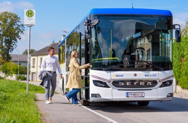 "leopoldi" der niederösterreichische Regionalverkehr, © NÖVOG/Bollwein Weißer Setra-Bus mit blauem Dach an Haltestelle, zwei Personen steigen ein, sonniger Tag, ländliche Umgebung.