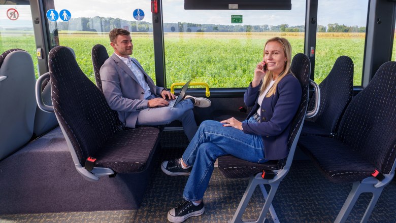 Zwei Personen sitzen sich in einem Bus gegen&uuml;ber, eine mit Laptop auf dem Scho&szlig;, die andere telefoniert, beide tragen Blazer und Jeans.