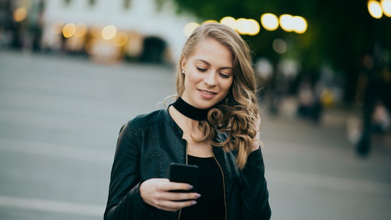 Junge Frau mit lockigem Haar und schwarzer Jacke liest l&auml;chelnd eine Nachricht auf ihrem Smartphone