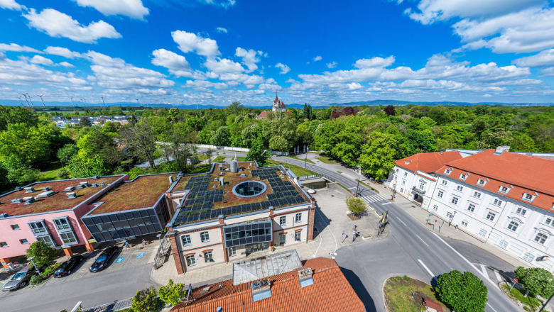 Luftaufnahme des Rathauses von Ebreichsdorf mit begrüntem Flachdach, umliegenden Straßen, Bäumen und einem roten Ziegeldach im Vordergrund.