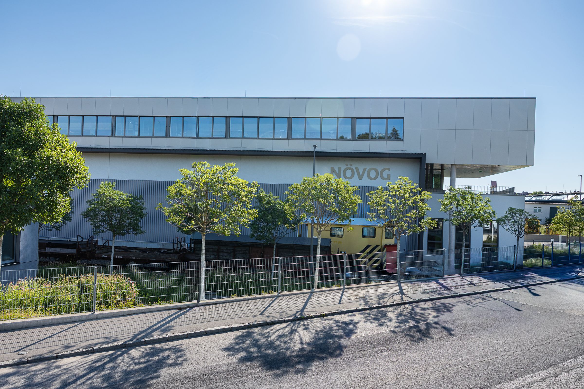 Modernes Gebäude mit langem Fensterband und gelbem Zugteil hinter Bäumen und Zaun an einer Straße