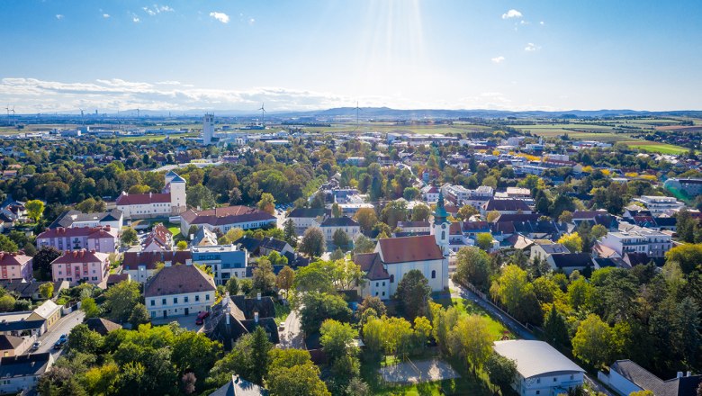Luftaufnahme einer Stadt mit vielen B&auml;umen, Wohnh&auml;usern und einem Kirchturm unter blauem Himmel mit vereinzelten Wolken.