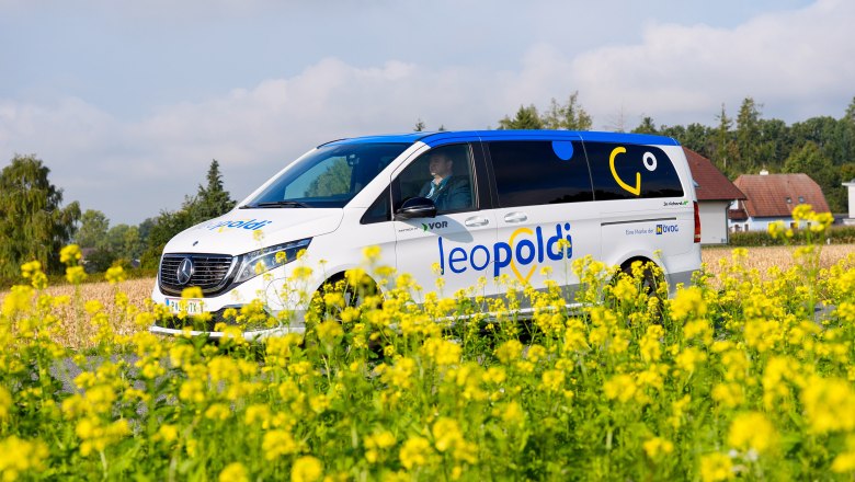 Weißer Mercedes-Van mit blauer und gelber Schrift 'leopoldi' auf der Seite, steht auf einem Feld mit gelben Blumen, im Hintergrund Bäume und ein Haus.