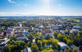 Luftaufnahme einer Stadt mit vielen B&auml;umen, Wohnh&auml;usern und einem Kirchturm unter blauem Himmel mit vereinzelten Wolken.